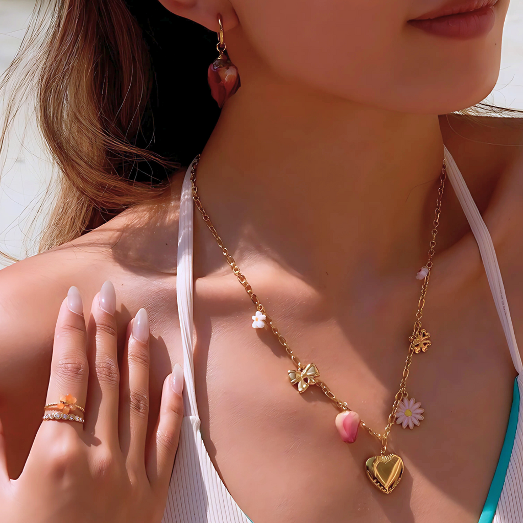 Woman wearing a gold charm necklace with heart and flower pendants, pink earrings, and rings.
