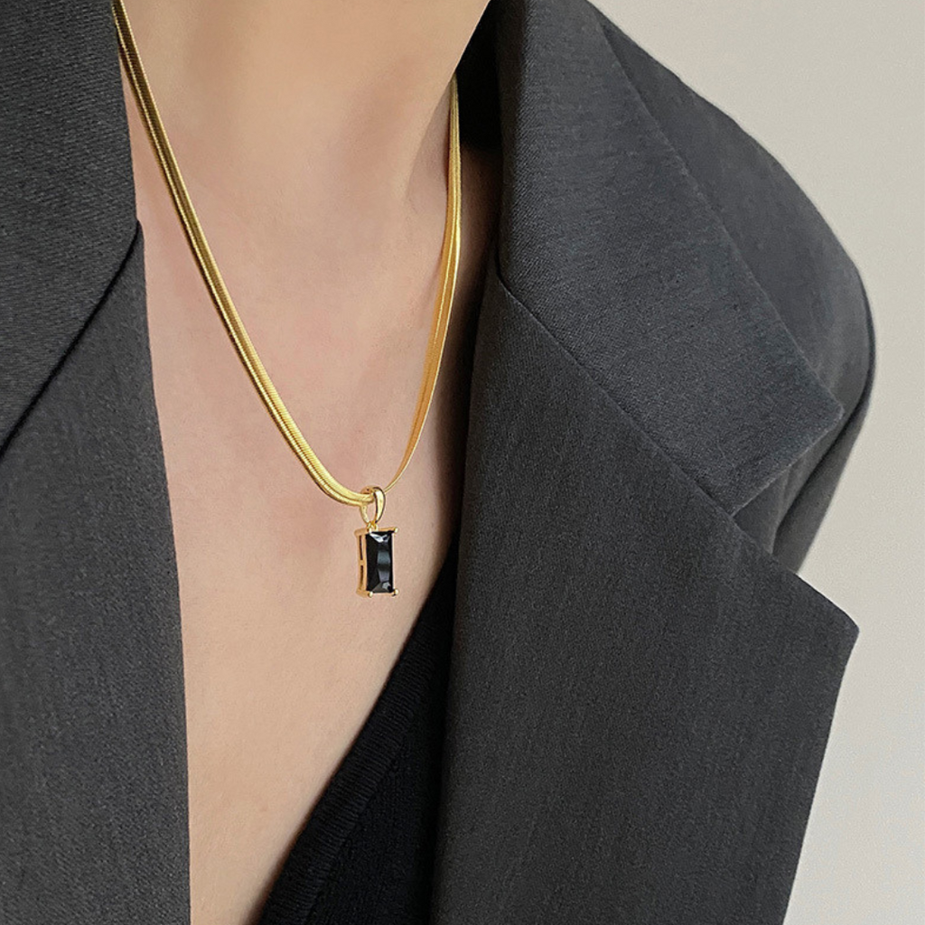 Gold necklace with a black pendant worn by a person in a gray blazer.